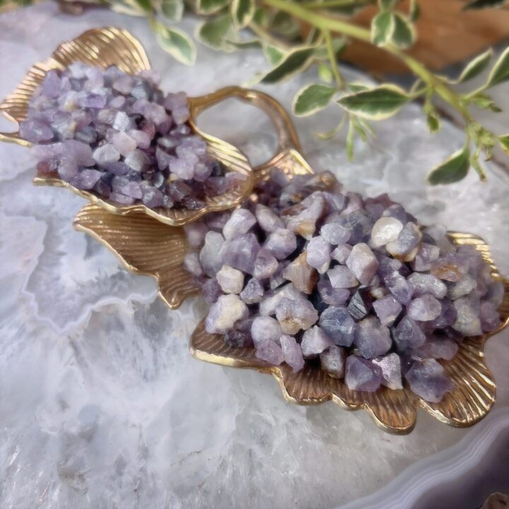 Raw purple gemstones displayed in gold shell-shaped holders on a light stone surface with soft natural greenery.